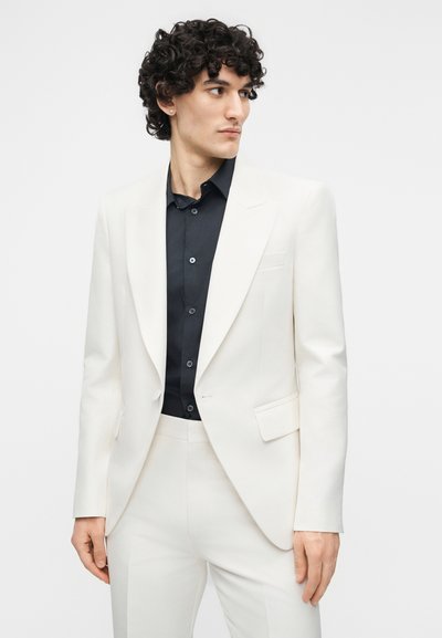 Άνδρας με σγουρά μαλλιά, που φοράει λευκό σακάκι και παντελόνι πάνω από μαύρο πουκάμισο με κουμπιά, κοιτάζοντας προς τα δεξιά μπροστά σε απλό φόντο.