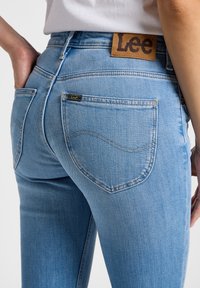 Persoană purtând blugi Lee albastru deschis, cu buzunare vizibile în spate, mâna sprijinită lângă șold și un top alb băgat în pantaloni.