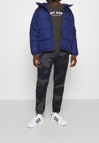 Blå puffjacka med huva, mörkgrå t-shirt, mörka byxor med kamouflage-mönster och vita sneakers med svarta ränder.