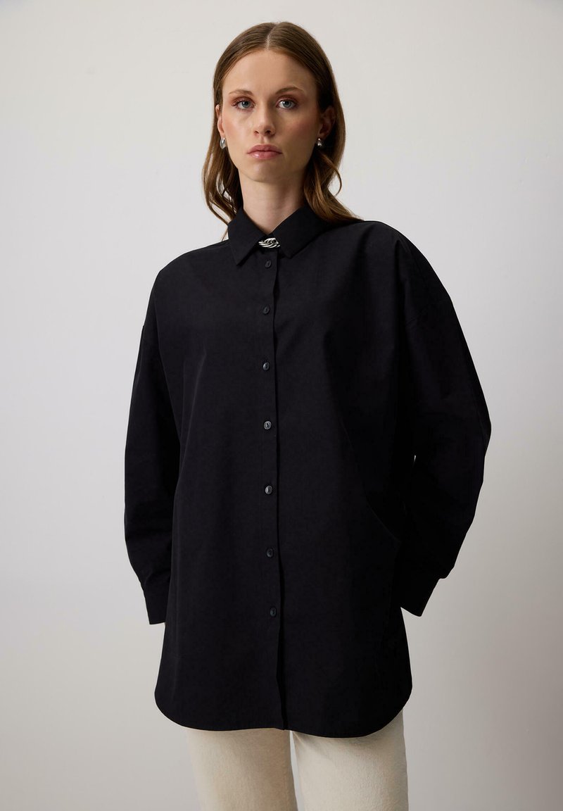 Giovane donna che indossa una camicia nera oversize con bottoni e maniche lunghe, abbinata a pantaloni color crema, davanti a uno sfondo chiaro e semplice.