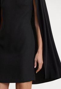 Svart kjole med figurformet liv og flytende cape-liknende ermer, laget av et glatt stoff med matt finish.