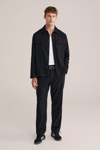 Svart jacka med två framfickor, bärs över en vit skjorta. Svarta byxor och svarta sneakers med vita detaljer. Slät tyg, skräddarsydd passform.