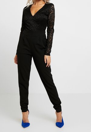 Zwarte kanten bodysuit met lange mouwen en een V-hals, gecombineerd met zwarte taps toelopende broek en blauwe puntige hakken. Gladde stof en een nauwsluitend ontwerp.