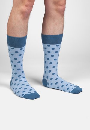 Nogi w jasnoniebieskich skarpetkach w ciemnoniebieskie groszki, palce, pięty i ściągacze w jednolitym ciemnoniebieskim kolorze, stojące na białej podłodze.