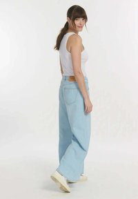  Lichtblauwe wijde jeans met een hoge taille, gestikte details en een bruine leren patch aan de achterkant. Draagt een witte tanktop.