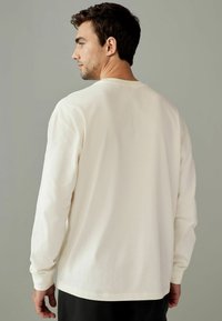 Long-sleeve cream shirt made from soft fabric, featuring a relaxed fit, round neckline, and ribbed cuffs, shown from the back.