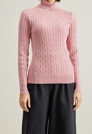 Femme portant un pull à col roulé moulant rose en maille torsadée et un pantalon noir ample sur un fond uni.