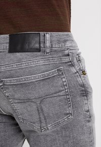 Primer plano de una persona que lleva jeans de mezclilla grises con un bolsillo trasero y una etiqueta de marca de cuero negro, combinados con una blusa marrón de canalé.