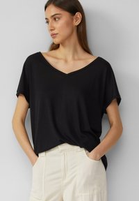 T-shirt noir à col en V, à manches courtes, en tissu doux. Présente une coupe décontractée et une fente sur les côtés ; associé à un pantalon de couleur claire.