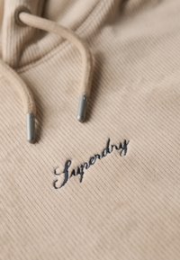 Felpa con cappuccio in corduroy beige caratterizzata da una superficie testurizzata, lacci con estremità metalliche argentate e un logo "Superdry" ricamato in blu scuro.