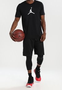 Svart t-shirt med vit Jordan-logga, svarta shorts, svarta kompressionsleggings och röda basket-skor, hållandes en basketboll.