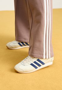 Zapatillas de crema con rayas de ante azul, cordones blancos y suela de goma. Combinadas con pantalones de chándal grises que presentan rayas laterales blancas.