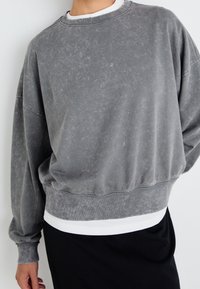 Persona vestida con un sudadera gris desgastada y holgada con puños y dobladillo acanalados, superpuesta a una camisa blanca y una falda o pantalones negros.