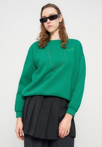 Πράσινο φούτερ με άνετη γραμμή, ριμπ μανσέτες και το κείμενο "BROOKLYN gallery" κεντημένο σε διακριτικό γκρι χρώμα. Συνδυασμένο με μια μαύρη πλισέ φούστα.