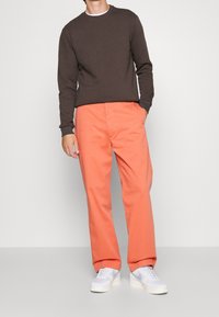 Suéter marrón de manga larga combinado con chinos naranjas y zapatillas blancas. El conjunto presenta un ajuste relajado y detalles de diseño simples.