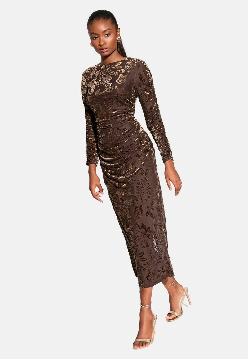 Langärmliges, tailliertes Kleid aus dunkelbraunem Samt mit goldfarbigem Blumenmuster, mit Rüschendetails und einem mittellangen Saum. Durchsichtige, hochhackige Sandalen.