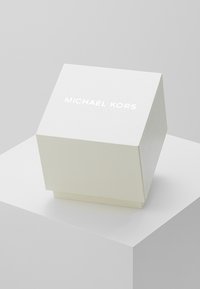 Scatola bianca a forma di piramide con finitura testurizzata, caratterizzata dal logo "MICHAEL KORS" in bianco sulla parte superiore. Bordo liscio e base resistente.