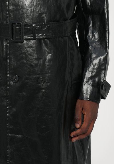 Abrigo negro de material brillante, con un diseño de doble botonadura, cintura ceñida con cinturón, dos botones decorativos y una superficie texturizada.