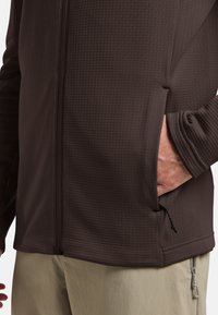 Veste zippée marron avec un motif texturé, dotée de poches latérales et de poignets élastiques. Le tissu semble lisse et léger.