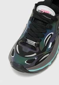 Zapatilla deportiva negra y verde con parte superior de malla, diseño texturizado, cordones negros y una etiqueta plateada de "Steve Madden" en la lengua.
