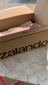 Pahvilaatikko, jossa on paksuilla mustilla kirjaimilla painettu "Zalando", sisältää ryttyistä raidallista kangasta ja se on asetettu marmoripinnalle.
