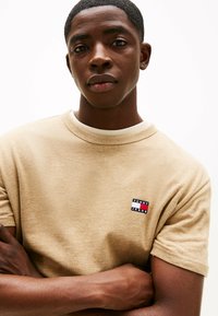 T-shirt beige a girocollo realizzato in un morbido tessuto, con un piccolo logo ricamato in rosso, bianco e blu nella parte sinistra del petto.