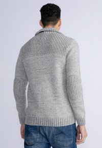 Pulover gri din tricot cu un guler șal cu nervuri, un corp texturat și pături la coate. Mâneca este de asemenea cu nervuri, iar croiala este lejeră.