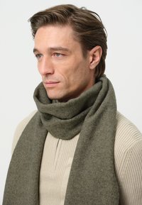 Sciarpa in lana verde oliva avvolta in modo casual intorno al collo, abbinata a un maglione a coste beige chiaro, che mette in risalto il contrasto di texture e colore.