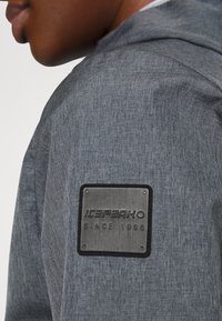 Haut à capuche en tissu gris texturé avec un patch métallique sur l'épaule portant le texte "ICEPEAK SINCE 1996". Design simple et coupe ajustée.