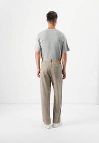 A person stands with their back to the camera, wearing a gray t-shirt and beige trousers, against a minimalist white background.