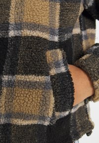 Fleece jacket with a plaid pattern in beige, gray, and black. Features textured fabric and a side pocket with a rolled cuff detail.
