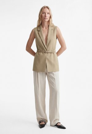 Frau trägt ärmellosen beigen Blazer mit Gürtel, weite cremefarbene Hosen und schwarze spitze Schuhe, steht mit verschränkten Händen im Rücken.