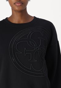 Melnā džemperī ir teksturēts, reljefs logo dizains centrā. Audumam ir gluda virsma, un tam ir ribota apkakle.