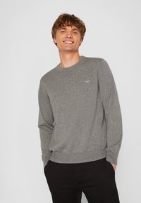 Smiling young man with curly hair wearing a grey crewneck sweater and black pants, standing with hands in pockets against a plain background.