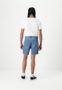 Shorts din denim de culoare albastru deschis, cu un tiv destrămat, asortați cu un tricou alb simplu. Vedere din spate, evidențiind cele două buzunare din spate.