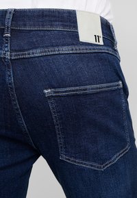 Tmavě modré džíny z denimu s hladkou texturou, s bílou značkovou štítkem na zadním pásu a standardními kapsami.