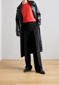 Trench-coat en cuir noir porté sur un pull rouge, associé à un pantalon noir taille haute et des chaussures noires ornées de clous.