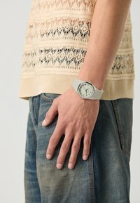 Montre gris clair avec un cadran arrondi, un design minimaliste et un bracelet en silicone. La main repose sur un tissu en denim avec un haut texturé en tricot.