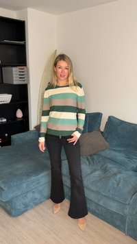 Kvinne som står ved en blågrønn sofa iført stripete genser, svarte utvide bukser og beige høyhælte sko i en minimalistisk stue.