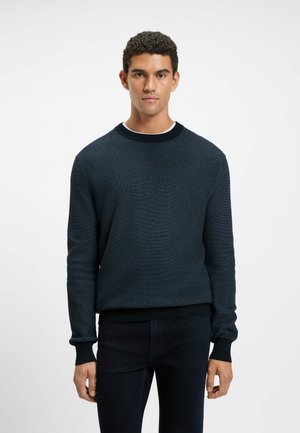 Marineblauwe gebreide trui met een ronde hals en geribbelde manchetten, gecombineerd met donkere jeans. De stof heeft een subtiel patroon en een comfortabele pasvorm.