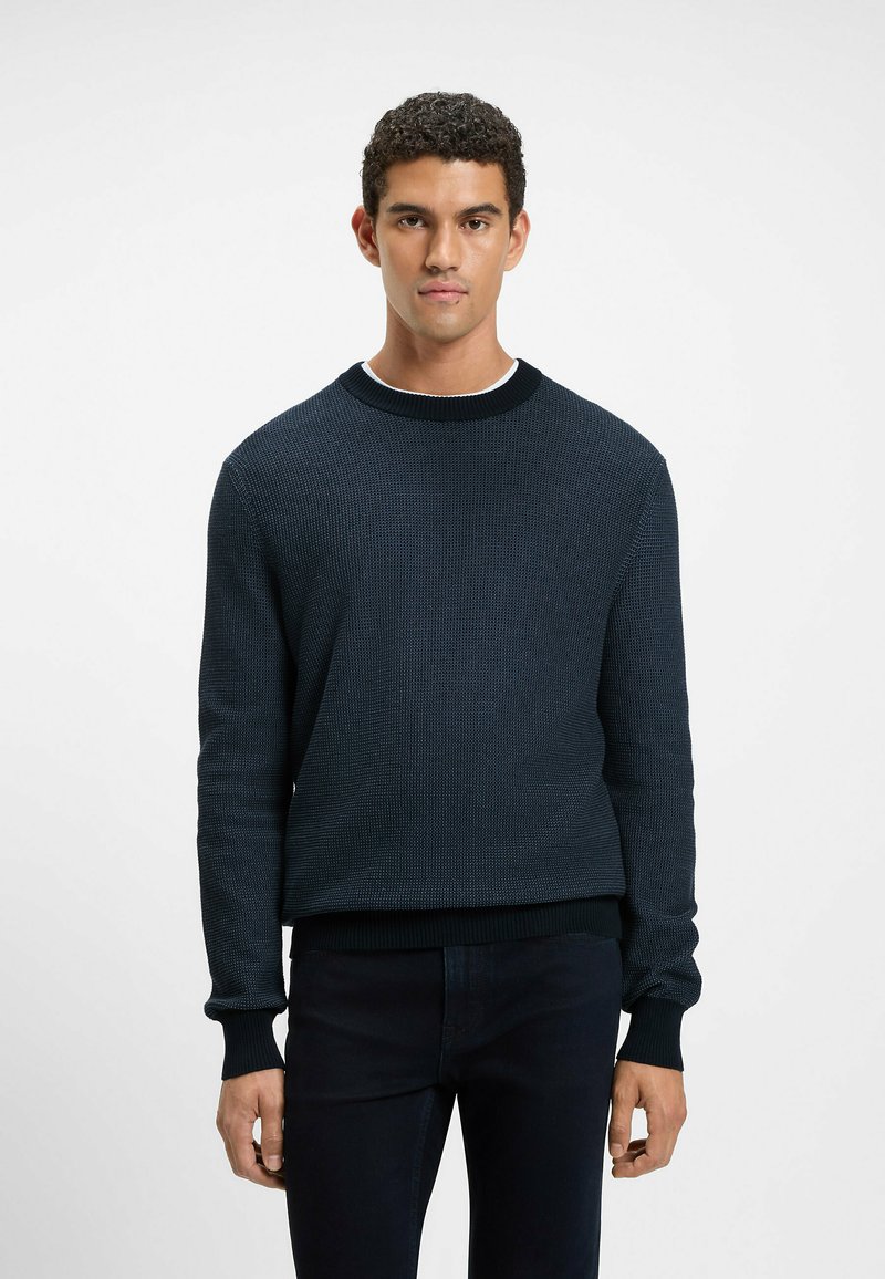 Navy textured knit sweater with a crew neck and ribbed cuffs, paired with dark jeans. The fabric has a subtle pattern and a relaxed fit.