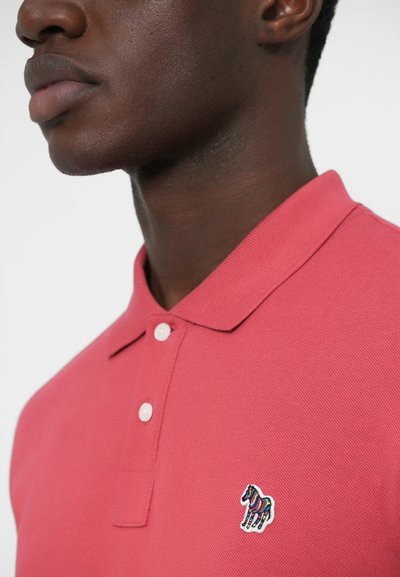 Punane polokleit tekstureeritud kangaga, millel on vasakul rinnal mitmevärviline sebra tikand ja valged nupud.