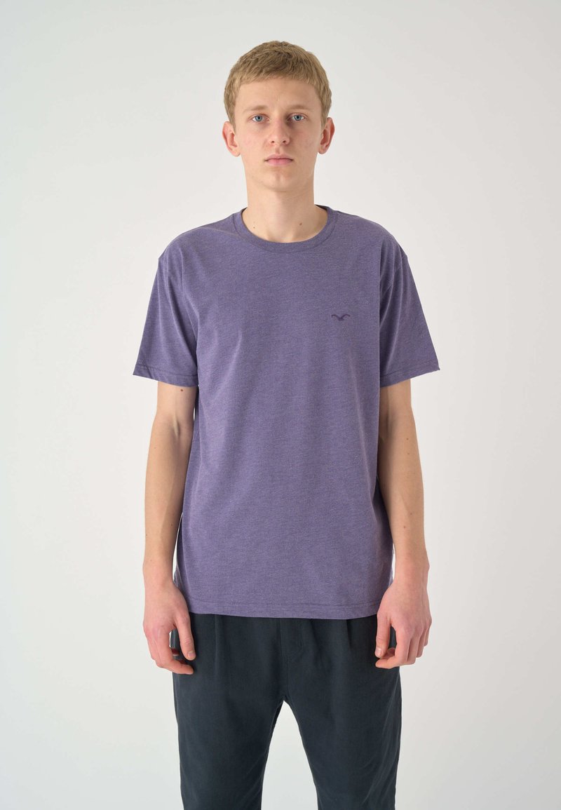 Kurzärmeliges lila T-Shirt aus weichem Stoff mit Rundhalsausschnitt und kleinem Logo auf der Brust, kombiniert mit dunklen, lockeren Hosen.