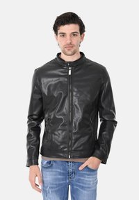 Giubbotto in finta pelle nera con colletto a scatto, chiusura con zip, due tasche laterali e una texture liscia. Indossato sopra una maglietta di colore chiaro.