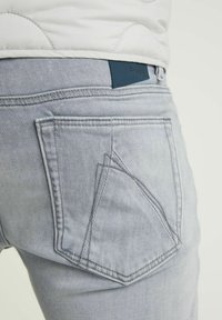 Světle šedé džíny z denimu s unikátním úhlovým designem zadní kapsy a malou modrou koženou značkovou nášivkou nad pasem.