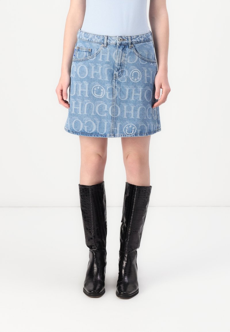 Jupe trapèze en denim avec des lettres blanches et des visages souriants sur toute la surface, portée avec des bottes noires texturées montantes et un haut blanc rentré dans la jupe.
