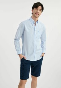Jasnoniebieska koszula w kratę z długimi rękawami, zestawiona z ciemnoniebieskimi szortami. Koszula ma przednią kieszeń i miękką fakturę materiału.
