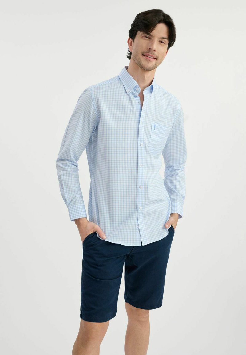 Jasnoniebieska koszula w kratę z długimi rękawami, zestawiona z ciemnoniebieskimi szortami. Koszula ma przednią kieszeń i miękką fakturę materiału.