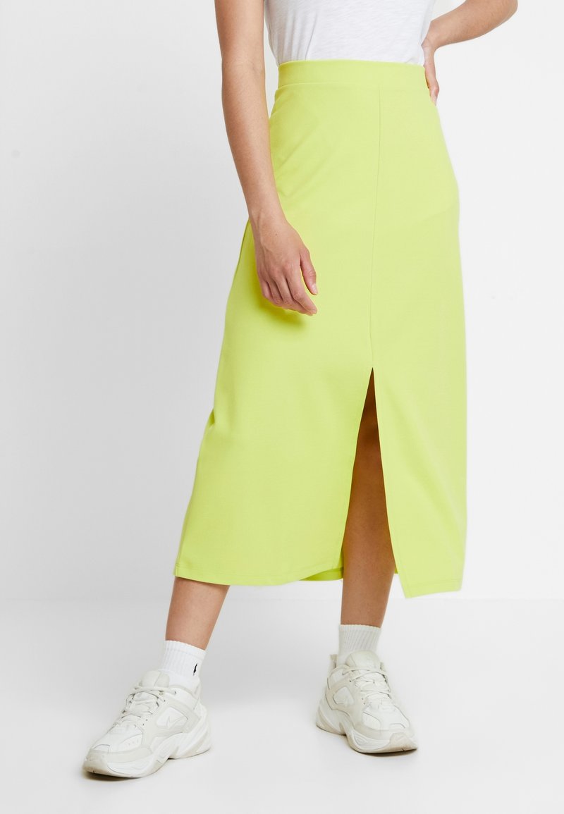 Jupe midi vert citron en tissu extensible avec une fente à l'avant, associée à des baskets blanches et des chaussettes hauteur cheville. Texture lisse et coupe ajustée.