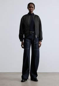 Crna bomber jakna s patentnim zatvaračem, nošena preko mornarsko plavog dolčevita. Labave tamne traperice s zakrpnim džepovima i kontrastnim šavovima. Crne cipele.